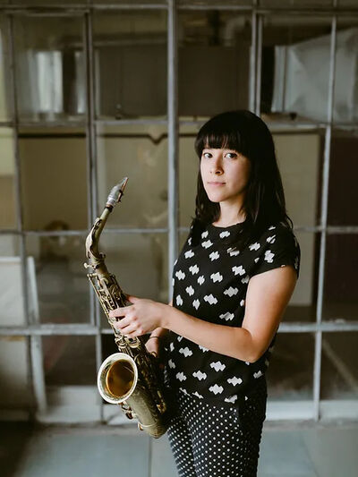 Person with bangs wearing a polka dot outfit, holding a saxophone while standing in an abandoned building.