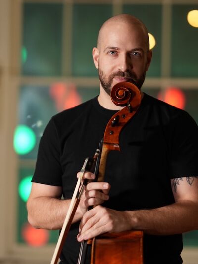 A bald person wearing a black t-shirt, holding a cello, in front of a wall with a window with blurred lights in the background.