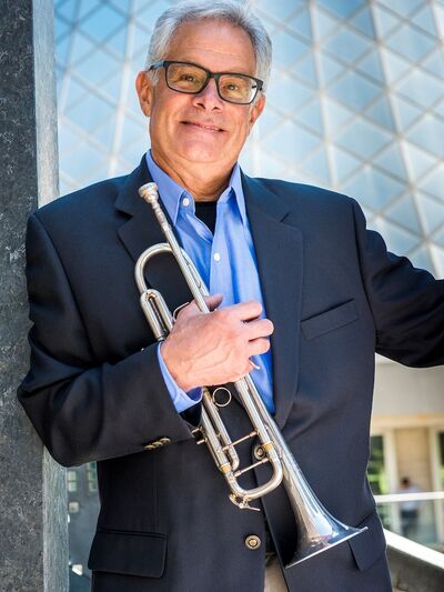 Person wearing a suit, holding a trumpet in front of a glass building.