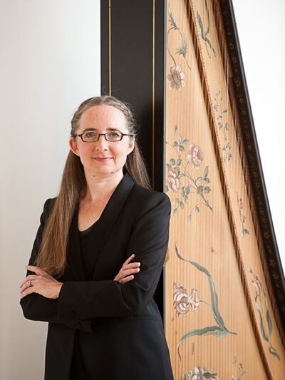Person wearing all black, with their hair half up, standing in front of a harp with their arms crossed, creating an elegant and poised look.
