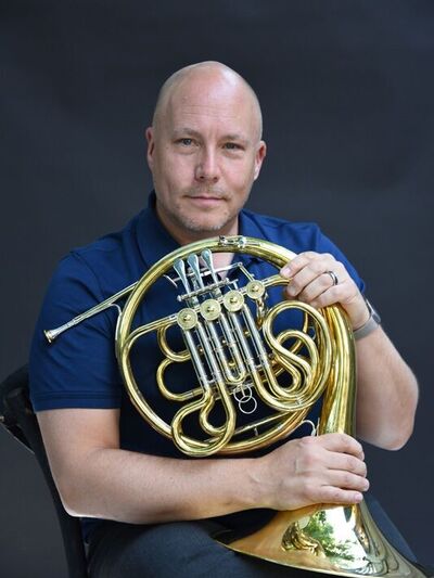 Headshot of a person with no hair in a blue polo shirt sitting in a chair holding a French horn with a dark grey background.