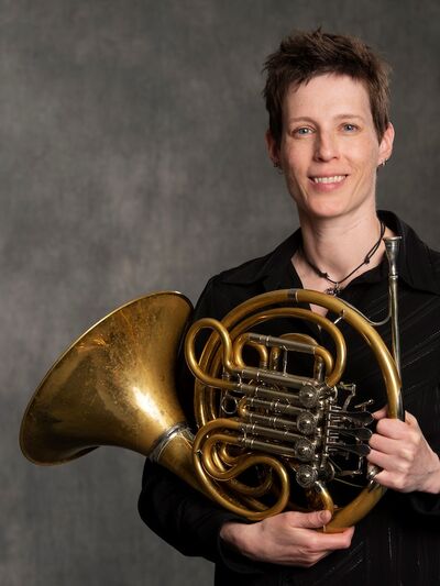 Person smiling, wearing a black shirt, holding a French horn in front of a grey background, creating a balanced and professional appearance.