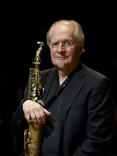 Headshot of a person wearing glasses and a suit, holding a saxophone.