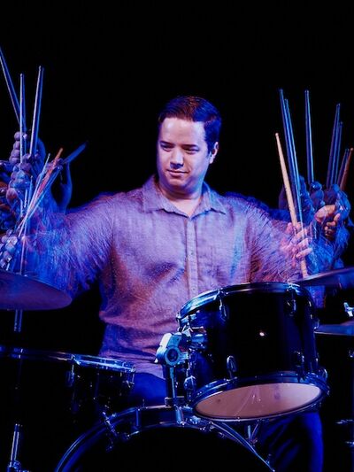 A person wearing a grey button-up shirt plays a drumset under blue and purple lighting, set against a black background.