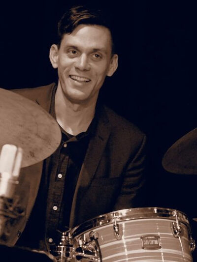 Black and white photo of a person wearing a button-up shirt and suit jacket, smiling while sitting in front of a drum set with a microphone.