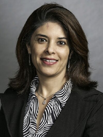 Headshot of a person wearing a striped shirt and blazer, smiling in front of a grey background.