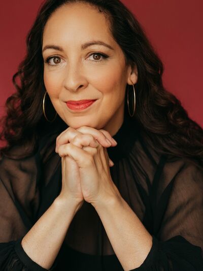 Headshot of a person wearing a black shirt and large hoop earrings, resting their head on their hand, with a bold red background adding contrast.