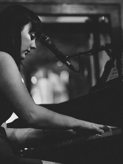 Black and white photo of a person playing the piano and singing with a microphone, with their attention focused on the keys.
