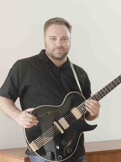 A person wearing a black short-sleeve button-up shirt is holding a guitar, leaning against a wooden cabinet in front of a white wall."