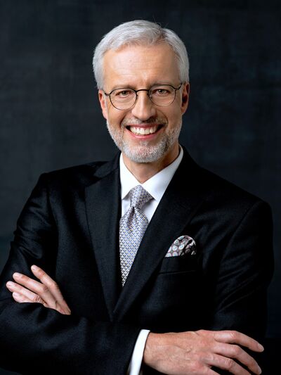 Person smiling, wearing a suit, standing in front of a dark background, exuding confidence and professionalism.