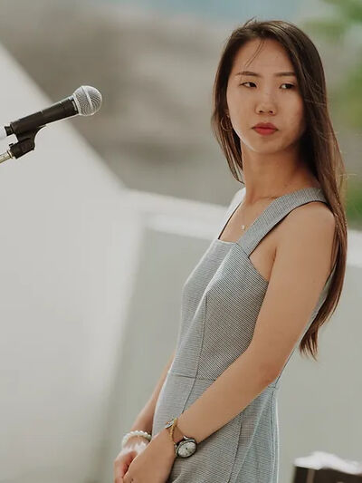 Person in a grey dress standing in front of a microphone, looking toward the side stage, possibly awaiting a cue or preparing for their next move.