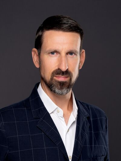 Headshot of a person wearing a blue suit, looking at the camera with a confident expression against a grey background.