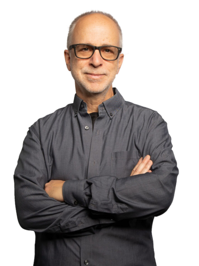 Man wearing glasses and a gray button-down shirt, standing in front of a white backdrop.