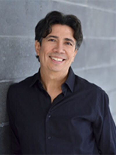 Person wearing a black button-down shirt, smiling in front of a grey brick wall.