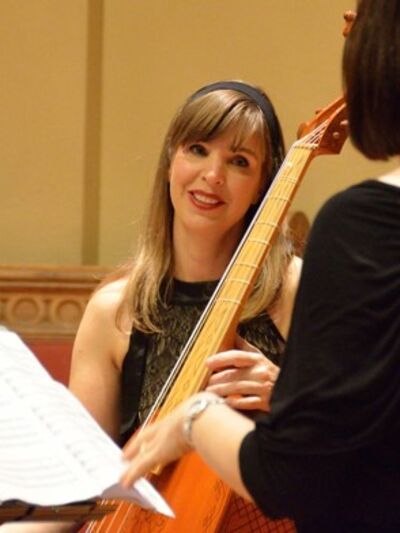Person holding a cello, attentively listening to the conductor's instructions, with the conductor and a music stand visible in the frame.