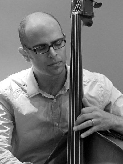 Black and white photo of a person wearing glasses and a button-up shirt, with their eyes closed, playing the double bass, capturing a moment of deep concentration and passion.