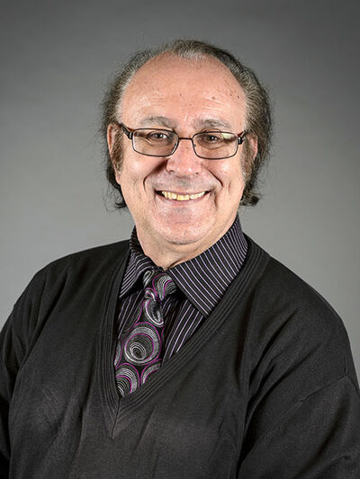 Headshot of a person wearing a black sweater vest and a purple tie with a circle pattern, smiling in front of a grey background.