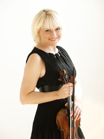 Person smiling while wearing a black dress, holding a violin and bow, and posing for the camera against a white background.