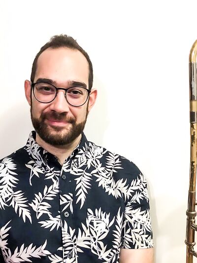 Person wearing a short-sleeve button-up shirt with a black-and-white leaf pattern, holding a trombone and smiling warmly.
