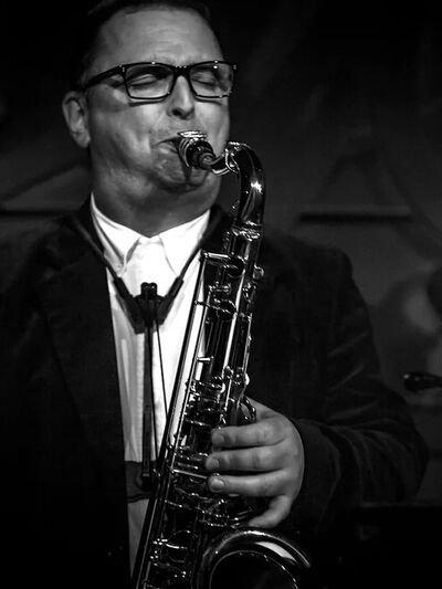 Black and white photo of a person wearing a suit and glasses, playing the tenor saxophone, captured in a moment of focused performance.