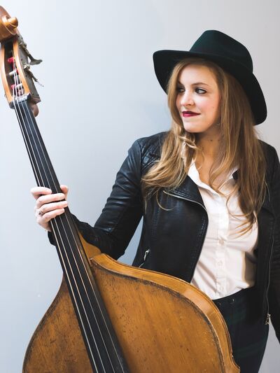 Person wearing a leather jacket and a cowboy hat, holding a double bass and looking to the side.
