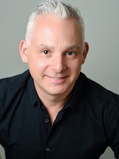 Headshot of a person wearing a black button-up shirt in front of a grey background, smiling.