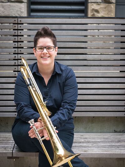 Person smiling, wearing all black, sitting on a bench with their legs crossed, holding a trombone.