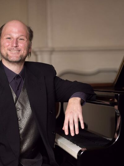 Person wearing a suit, smiling, and leaning on a grand piano.