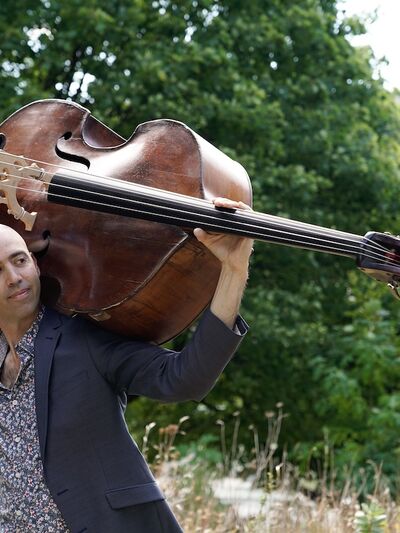 Person wearing a suit, holding a double bass behind their head in a field with trees in the background.
