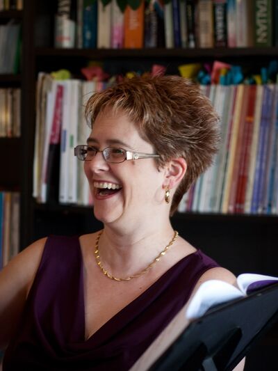 Person with short hair, wearing a purple dress, laughing while holding a book in a library.