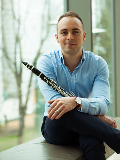 Person sitting down in front of a wall of windows, wearing business clothes and holding a clarinet.