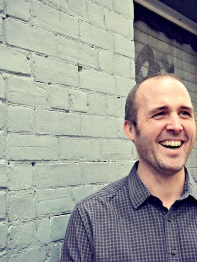 Headshot of a person smiling joyfully, wearing a grey button-down shirt, standing in front of a grey painted brick wall.