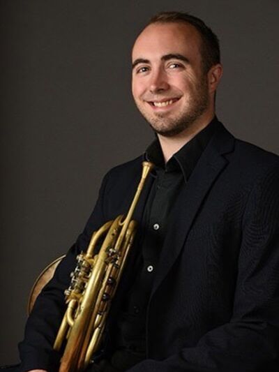 Headshot of a person smiling, wearing a black suit, holding a trumpet, sitting in a chair with a grey background.