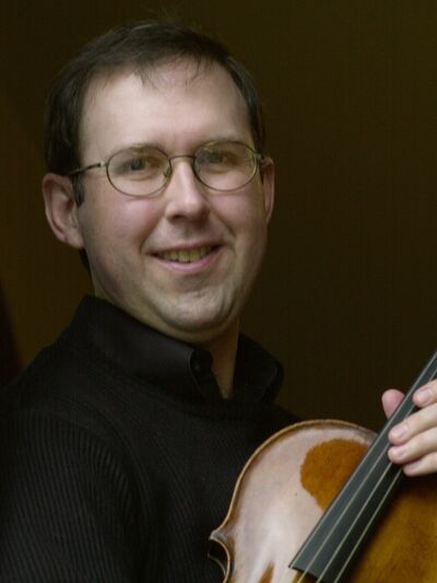 Person smiling while wearing a black shirt, holding a viola in front of a brown background.