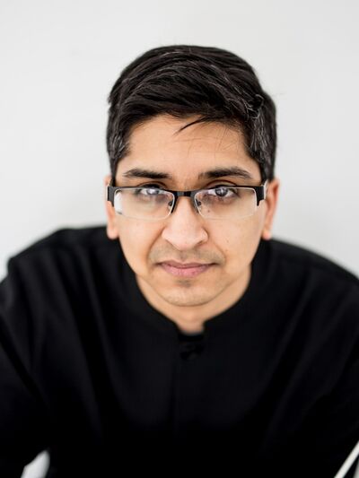 Headshot of a person wearing glasses and a black sweater, leaning slightly and holding a conductor's wand, with a light grey background.