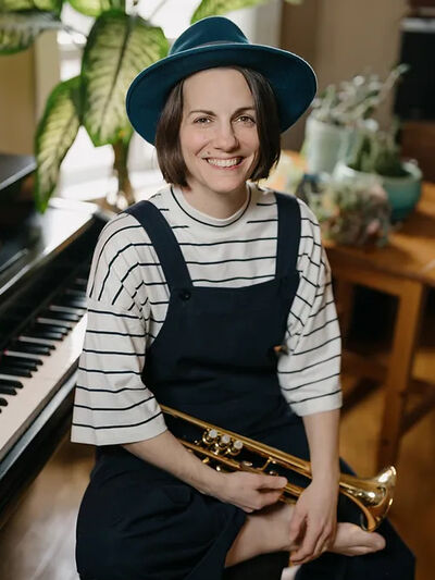Person wearing a blue hat and black dress, holding a trumpet while sitting on a piano stool in front of a piano, with plants surrounding the scene, creating a serene and artistic atmosphere.