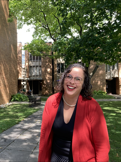 Person wearing glasses and a red jacket smiling. Standing on grass in front of tree and building