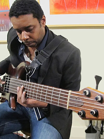 Person in a suit jacket and jeans playing an electric bass in front of a backdrop of colorful paintings.