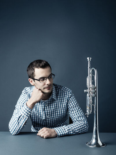 Person wearing a blue and white checkered shirt, with their hand on their chin, looking at a trumpet standing on the table. Both the background and the table are grey.