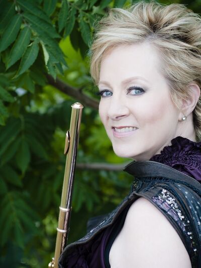 Headshot of a person looking over their shoulder, wearing black and holding a flute, with greenery in the background.