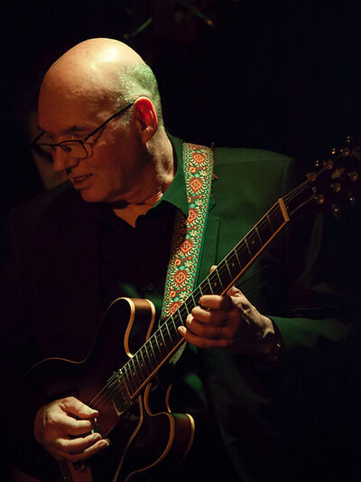 Person wearing all black playing the guitar, looking into the audience, illuminated by stage lights.