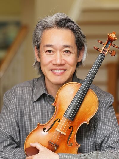 Person wearing a grey shirt, smiling while holding a violin.