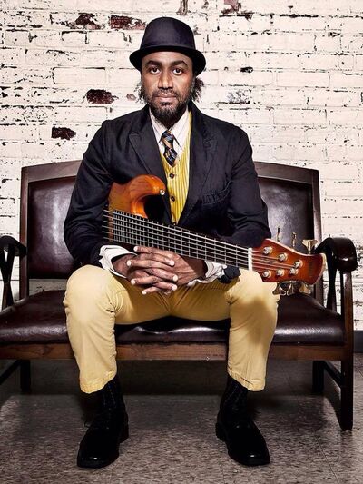 A person wearing a black and yellow suit with a black fedora, sitting on a bench with their fingers interlaced, holding a guitar in front of a white painted brick wall.