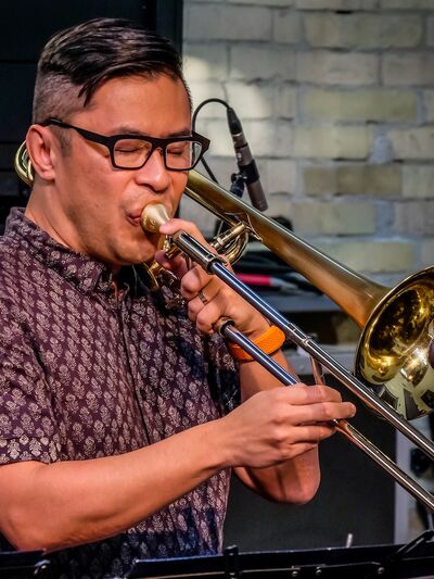 A person with glasses, wearing a red patterned short-sleeve button-up shirt, is playing the trombone while surrounded by sound equipment.