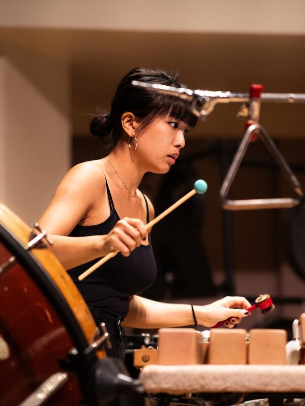Percussionist wearing black, playing instruments and focused on their sheet music.