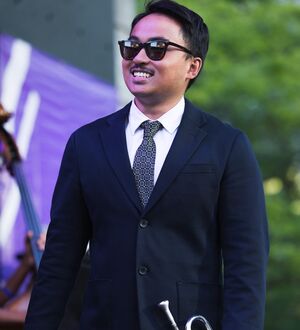 Person in a suit and tie wearing sunglasses, standing on a stage holding a trumpet