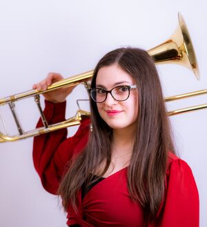 Person with long hair wearing a red shirt, holding a trombone on their shoulder.