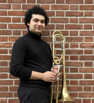 Headshot of a person wearing all black, holding a trombone against a brick wall.