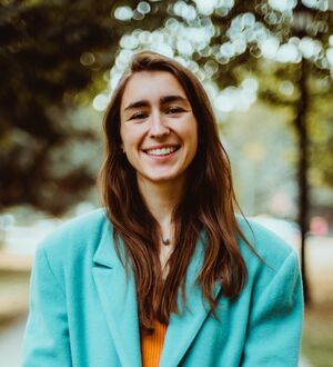 Headshot of a person wearing a teal blazer, smiling warmly while standing on a wooded path, with natural surroundings in the background.
