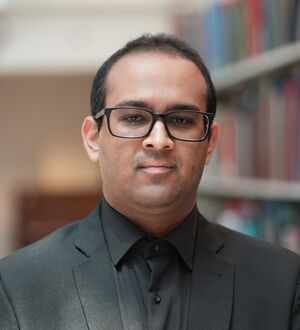 Headshot of a person wearing glasses and a black suit, standing in front of a bookshelf.